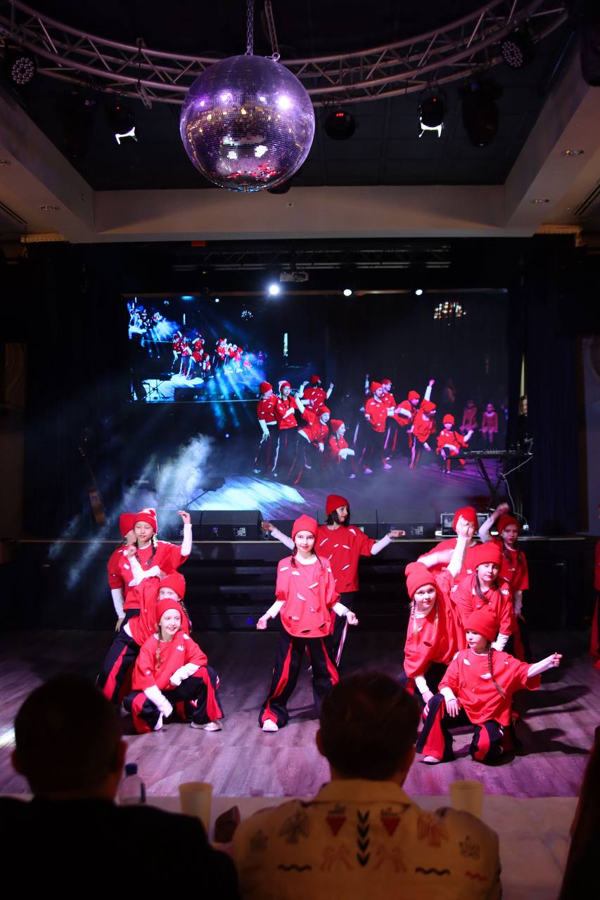 A dance group performing on a lit stage in front of a full hall
